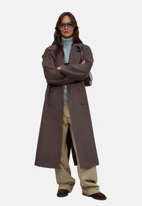 Długi, brązowy wełniany płaszcz o podwójnym zapięciu, noszony na jasnoniebieskim swetrze i beżowych spodniach z szerokimi nogawkami. Ciemne obuwie dopełnia stylizację.