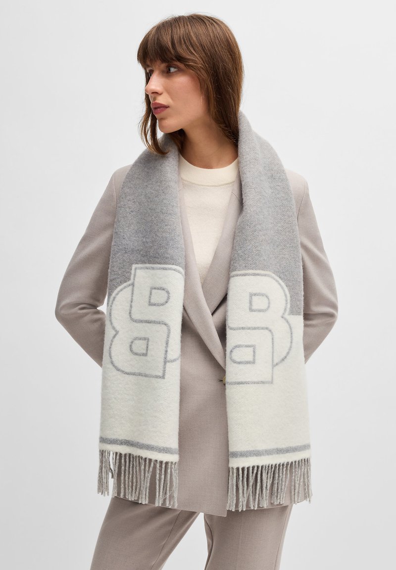 Écharpe en laine gris et blanc cassé avec un grand logo, texture douce et bords frangés, portée sur un pull beige et un costume.