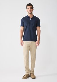 Marineblauwe polo met een kraag en een borstzak, gecombineerd met lichtbeige pantalons en bruine instappers. Glad materiaal, comfortabele pasvorm.