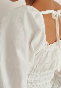 Kobieta ubrana w białą bluzkę z bufkami, marszczonym tyłem i zawiązanym kokardą na górze pleców.