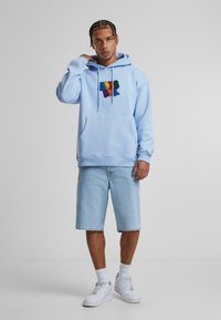 Helle blaue Hoodie aus Baumwolle, mit einem bunten Grafiklogo auf der Brust, kombiniert mit hellen Jeansshorts und weißen Sneakers.