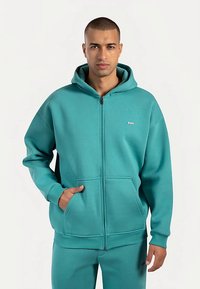 Sudadera con capucha color azul verdoso con cremallera, hecha de tela suave. Incluye capucha, bolsillos delanteros, puños acanalados y un pequeño logo en el pecho.