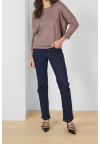 Pullover marrone a coste con maniche ampie, abbinato a jeans blu scuro e décolleté nere con punta appuntita e dettagli in fascia. Le calzature hanno una finitura lucida.