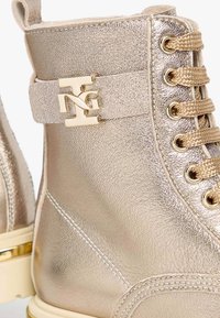 Metallisch goldfarbene Knöchelstiefel mit strukturiertem Lederfinish, goldenen Hardware-Akzenten und beigen Schnürsenkeln. Sie verfügen über einen gebrandeten Riemen und eine kontrastierende Sohle.