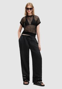 Zwart doorschijnend gebreid topje met korte mouwen, gecombineerd met losse zwarte broek met een trekkoord in de taille en decoratieve accenten aan de uiteinden. Zwarte sandalen.