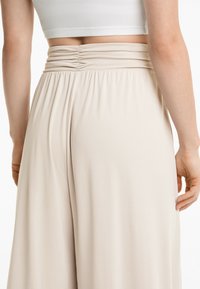 Beige weit geschnittene Hosen mit weicher Textur und gerafftem Taillendetail. Der Stoff fällt fließend und zeigt ein glattes, minimalistisches Design.