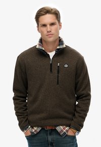 Bruine fleece met rits en een geruite kraag, textuurmateriaal, voorzak met rits en logo patch op de borst, in combinatie met een geruit shirt eronder.