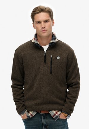 Brown zip-up fleece with a plaid collar, textured material, front zipper pocket, and logo patch on the chest, paired with a plaid shirt underneath.