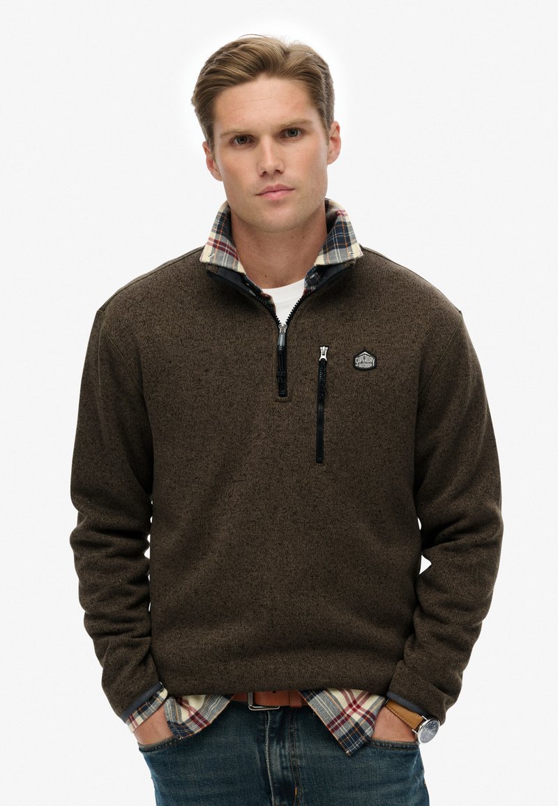 Bruine fleece met rits en een geruite kraag, textuurmateriaal, voorzak met rits en logo patch op de borst, in combinatie met een geruit shirt eronder.