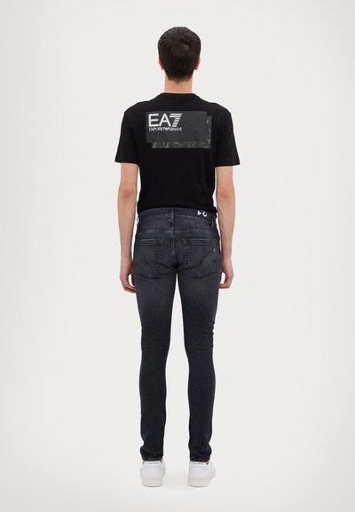 T-shirt nera con grafiche bianche che riportano "EA7 EMPORIO ARMANI" sulla schiena, abbinata a jeans scuri e sneakers bianche.