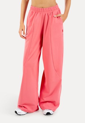 Osoba nosząca luźne różowe spodnie z elastycznym pasem i białe trampki, stoi z jedną ręką w kieszeni.