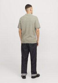 Polo de punto en verde apagado, de mangas cortas; combinado con unos pantalones de mezclilla oscuros, con los puños remangados y zapatos negros, visto desde la parte posterior.