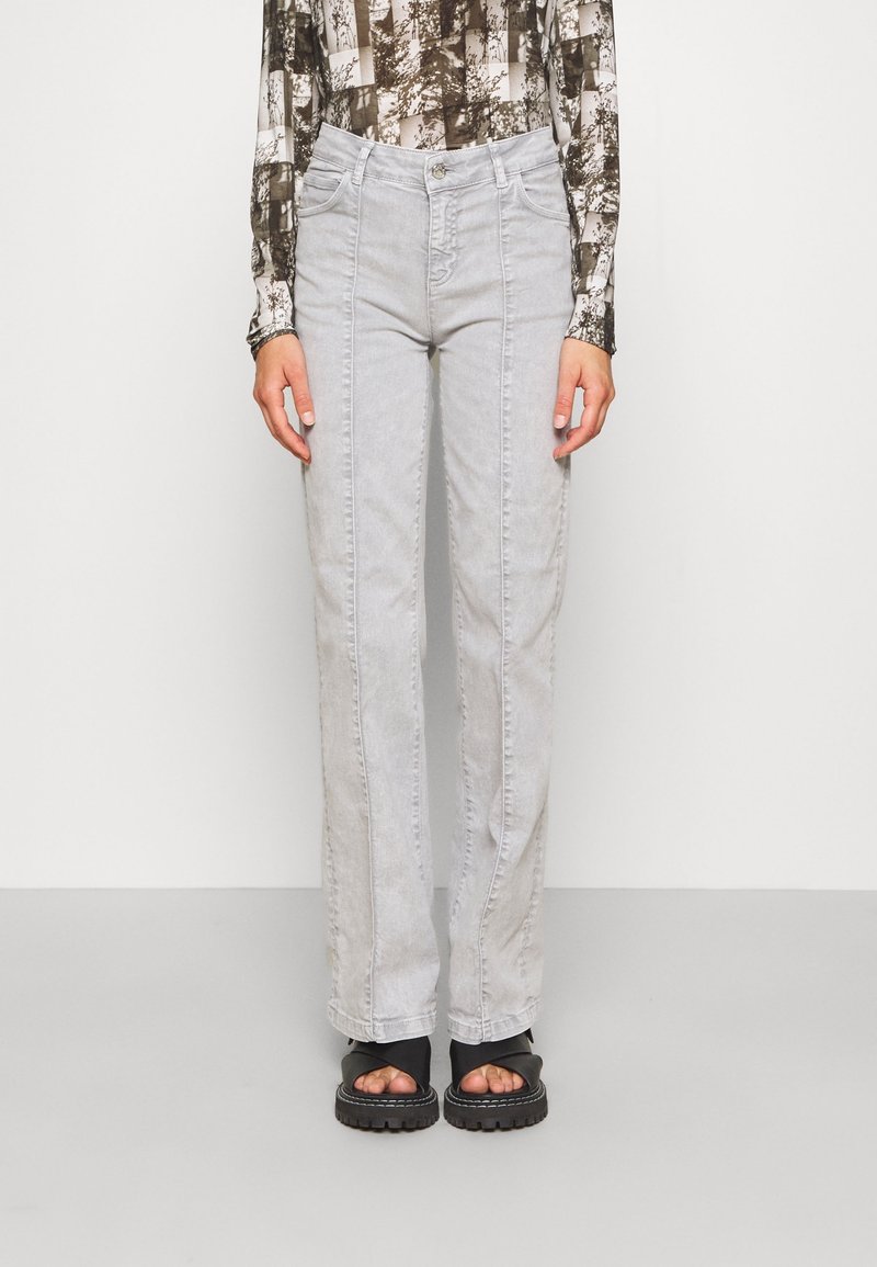 Personne portant un jean taille haute gris clair avec des coutures visibles le long des jambes, associé à des sandales noires épaisses et un haut à manches longues à motifs.