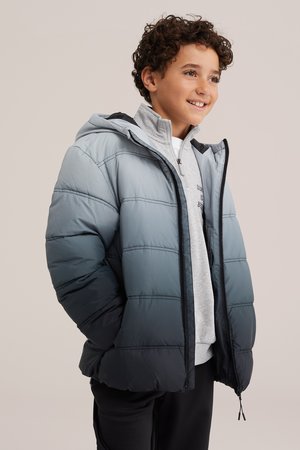 Garçon souriant aux cheveux bouclés porte une doudoune matelassée bleu clair par-dessus un sweat-shirt gris devant un fond uni.