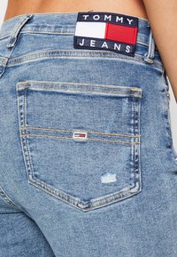Jeans de denim de color azul claro con un bolsillo trasero que presenta un pequeño parche de logo en rojo, blanco y azul marino. Incluye un detalle desgastado en la tela.