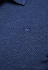 Tamsiai mėlyna polo marškinėliai, pagaminti iš tekstūruotos medžiagos; ant krūtinės yra siuvinėtas arkliuko logotipas. Klasikinio dizaino apykaklė su trumpomis rankovėmis.