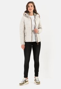 Lichte, romige pufferjack met horizontale quiltstiksels, een capuchon en ritssluiting. Gecombineerd met zwarte jeans en multikleurige sneakers.