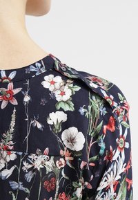 Marinblå bluse med blomsterprint, der har en knapdetalje ved skulderen, med livlige blomster, blade og en glat, blød tekstur.