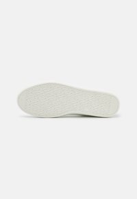 Schuhsohle, weißes Gummi mit eingeprägtem Logo-Muster. Flache Form, abgerundete Spitze, strukturiert für Grip. Glatte Kanten, minimalistisches Design.