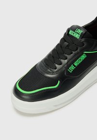Zwarte sneaker van mesh en leer. Heeft groene accenten, een ronde neus en een vetersluiting. Witte zool. Merkenlogo zichtbaar.