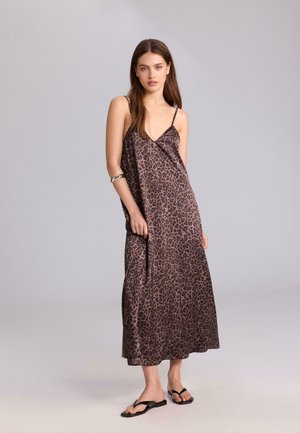 Junge Frau trägt ein ärmelloses, braunes Maxikleid mit Leopardenmuster und schwarze Flip-Flops, steht mit einer Hand auf dem Oberschenkel.