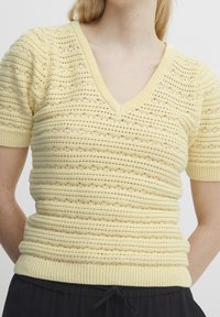 Gul strikket sweater med V-hals, korte ærmer og strukturerede striber. Har en tætsiddende talje og ribbede manchetter ved ærmerne og nederste kant.