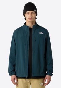 Teal soft-shell jacket with a zip front, high collar, and two side pockets. Logo on the upper left side. Worn over a black shirt and beige pants.