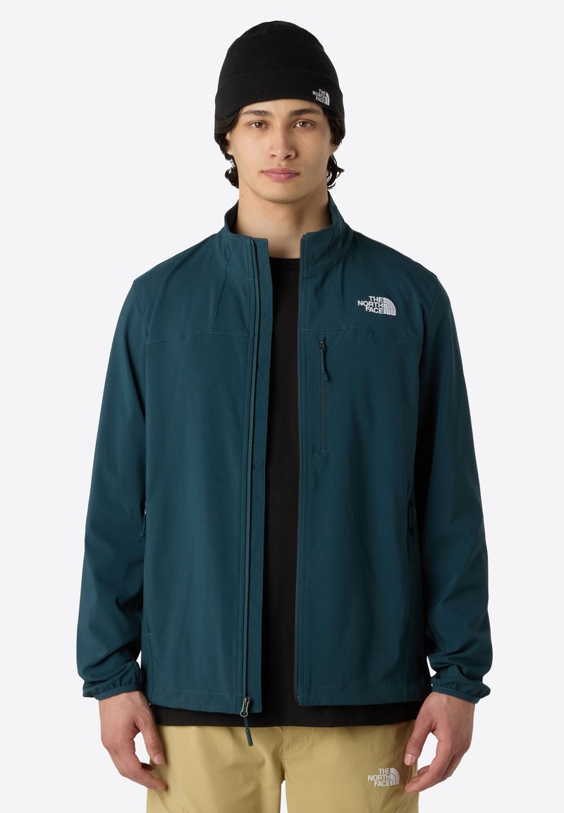 Teal soft-shell jacket with a zip front, high collar, and two side pockets. Logo on the upper left side. Worn over a black shirt and beige pants.