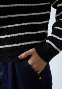 Svart- och vitrandig stickad tröja med ribbad nederkant, med snäv passform och en hand i en blå denimficka med synlig sömnad.