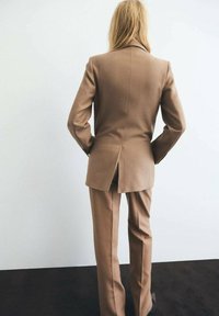 Traje marrón con una chaqueta ajustada y pantalones de pierna recta. La chaqueta presenta una abertura en la parte trasera, diseño a medida y textura suave.