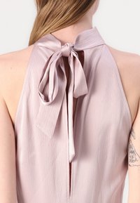 Ljust rosa ärmlös topp med hög krage och en knytåtta bak, tillverkad av textil med en subtil glans.