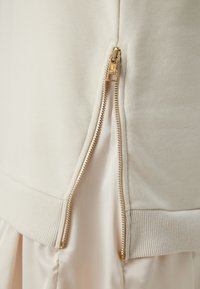 Indumento superiore con zip in tessuto color crema; presenta accessori in metallo dorato e orlo a costine testurizzate; tessuto interno simile alla satinato visibile.