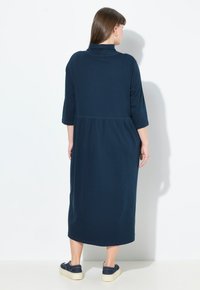 Tummansininen, pitkä mekko korkean kauluksen ja kolme neljäsosaa hihojen kanssa. Pehmeä kangas, jossa on laskostettu vyötärö, yhdistettynä rentoihin kenkiin.