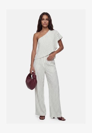 Femme portant un top blanc asymétrique et un pantalon large assorti à rayures verticales noires, tenant un sac à main tissé bordeaux.
