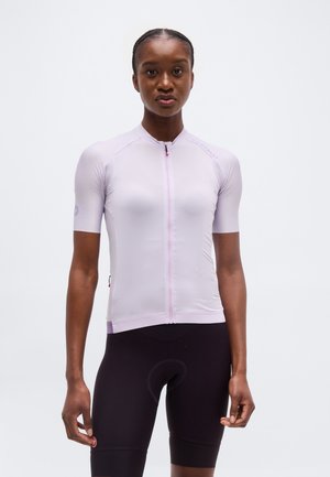Femme debout portant un maillot de cyclisme à manches courtes violet clair et un short de cyclisme rembourré noir, sur un fond uni.