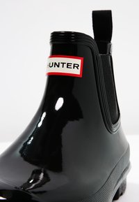 Czarny gumowy botek na kostkę o błyszczącym wykończeniu, z elastycznym bokiem oraz białą metką z czerwonymi akcentami z nazwą marki "HUNTER".
