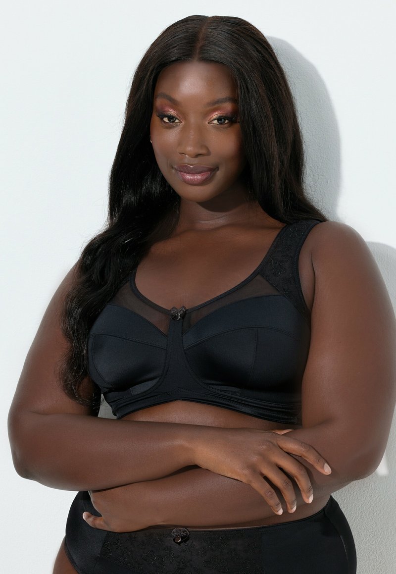 Reggiseno nero con inserti in rete trasparente, spalline larghe con dettagli in pizzo e un bottone decorativo al centro. Tessuto liscio e design di sostegno.