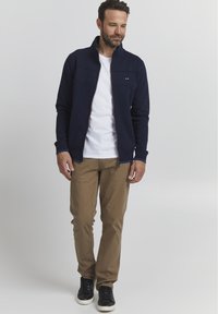 Giacca zip-up blu navy con colletto alto, t-shirt bianca sotto, pantaloni beige e sneakers neri. Presenta cuciture a contrasto sulla giacca.
