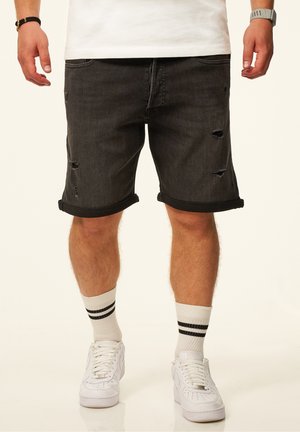 Mann trägt schwarze hochgekrempelte Jeansshorts mit Rissen, weiße Socken mit schwarzen Streifen und weiße Sneaker vor schlichtem Hintergrund.