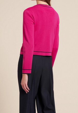 Cardigan lavorato a maglia fucsia con maniche lunghe, caratterizzato da una striscia nera lungo il collo e l'orlo, abbinato a pantaloni larghi neri.