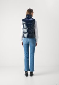 Gilet imbottito navy con finitura lucida, colletto alto, cuciture orizzontali, indossato sopra una camicia bianca, abbinato a jeans svasati azzurri chiari e stivali neri.