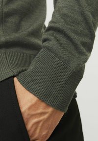 Mânica unui pulover verde tricotat, cu textură în relief, care prezintă un design ajustat. O mână este parțial vizibilă, sprijinindu-se pe un material negru.