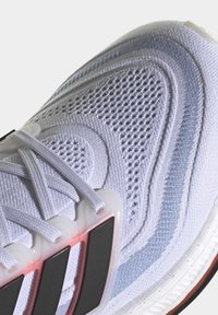 Gros plan d'une chaussure de sport blanche en tissu mesh, motifs texturés, lacets blancs, et design rayé noir près de la semelle.
