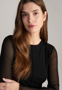 Schwarzes Kleid mit langen, durchsichtigen Ärmeln, glatter Textur und tailliertem Design. Das Model hat schokoladenbraunes Haar und einen neutralen Hintergrund.
