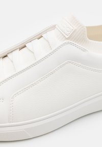 Vit sneaker med ett texturerat läderöverkast, elastiska snören och en slät gummisula. Har en subtil chevrondesign på sidan.
