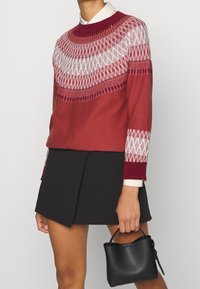 Czerwony sweter z białym geometrycznym wzorem na dekolcie i rękawach, w połączeniu z czarną spódnicą, trzymany w małej czarnej torebce.