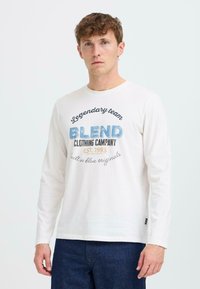 Hosszú ujjú fehér pamut póló, kék és barna szövegnyomattal. A felirat: "Legendás csapat BLEND Clothing Company ALAPÍTVA 1993."