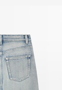 Vue arrière d'un jean en denim bleu clair montrant une passants de ceinture, une ceinture, et une seule poche arrière avec des coutures visibles.