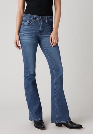 Kobieta w niebieskich dżinsach bootcut z wysokim stanem i czarnych botkach na obcasie, stojąca na tle jasnego, jednolitego tła.