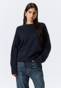 Pull bleu marine, surdimensionné, fabriqu�é à partir d'un matériau doux et duveteux, avec un col rond et de longues manches légèrement larges. Porté avec un jean.